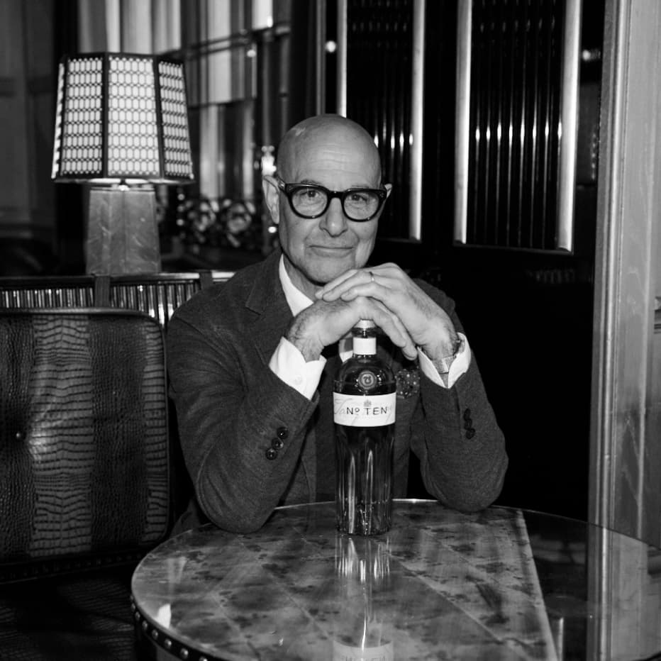 Stanley Tucci sitting a tabled with his arms folded around a bottle of Tanqueray No. Ten