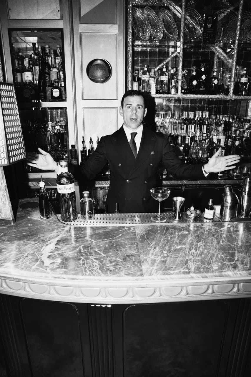A black and white image of a man standing behind a bar. On the bar in front of him is an array of cocktail making equipment and a bottle of Tanqueray No. TEN.