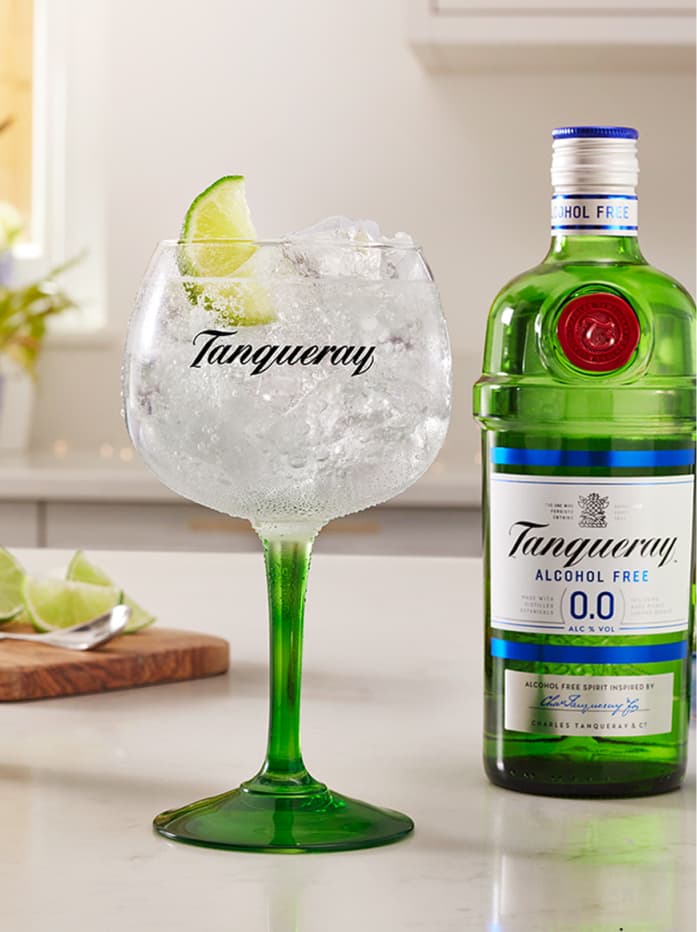 A Tanqueray 0.0 & Tonic cocktail garnished with a lime wedge on a table with a bottle of Tanqueray Alcohol Free 0.0 sitting behind the cocktail