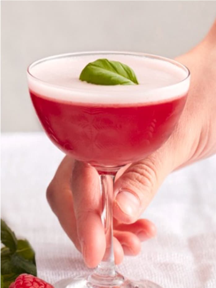 A hand holding a red Tanqueray Royale Clover cocktail garnished with a mint leaf.