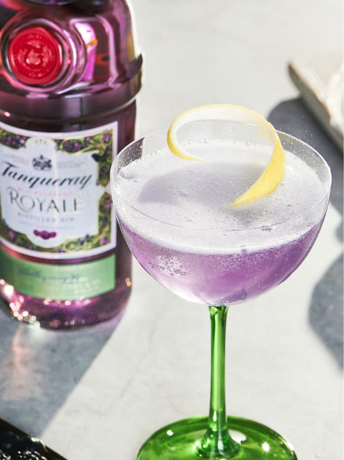 A purple Tanqueray Blackcurrant Royale French 75 cocktail in a glass garnished with a lemon swirl. There is a bottle of Tanqueray Blackcurrant Royale in the background.