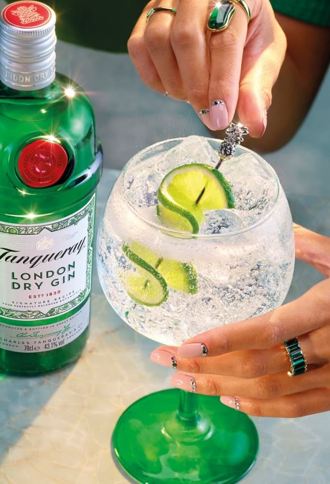 A Gin & Tonic cocktail made with Tanqueray London Dry Gin garnished with two lime slices. A bottle of Tanqueray London Dry gin sits in the background of the image.